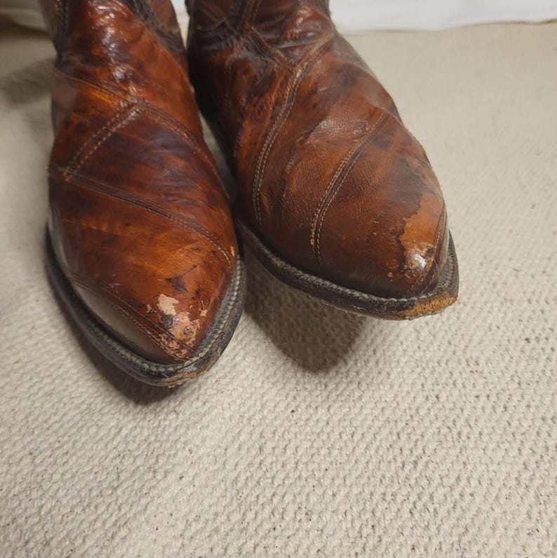 May include: A pair of well-worn brown leather cowboy boots. The boots have a pointed toe and a stitched design. The leather shows signs of wear and tear, with some scuffing and discoloration. The boots are set against a neutral background.