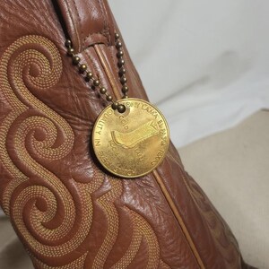 May include: Brown leather cowboy boot with intricate stitching and a gold-colored tag. The tag reads "Tony Lama El Paso, Texas" and features a boot illustration. The boot has a decorative stitched pattern.