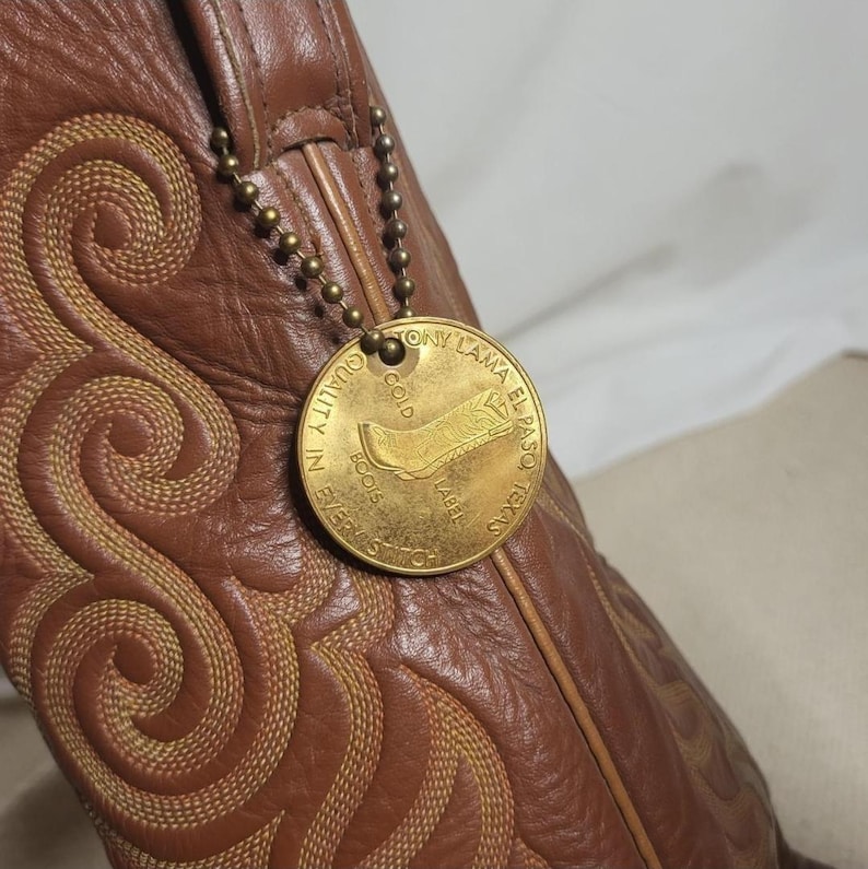 May include: Brown leather cowboy boot with intricate stitching and a gold-colored tag. The tag reads "Tony Lama El Paso, Texas" and features a boot illustration. The boot has a decorative stitched pattern.