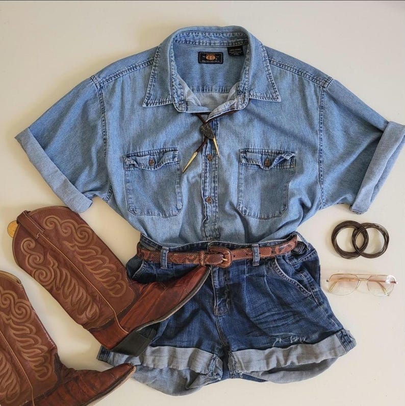 May include: A denim shirt with rolled-up sleeves, denim shorts, and brown leather cowboy boots. A brown leather belt, two dark brown hair ties, and a pair of glasses complete the outfit. A bolo tie is also included.