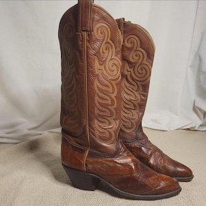 May include: A pair of brown leather cowboy boots. The boots have intricate stitching and a pointed toe. The leather is a rich, warm brown color with darker accents. The boots have a classic Western design.