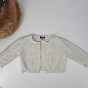 May include: A cream-colored, long-sleeved knit cardigan with a round neckline and an open front design. The garment features ribbed cuffs and a ribbed hemline. A black brand label with white text is sewn into the interior of the neckline.