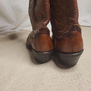 May include: A pair of brown leather cowboy boots. The boots have decorative stitching and a pointed toe. The heels are dark brown and angled. The boots are on a light-colored surface.
