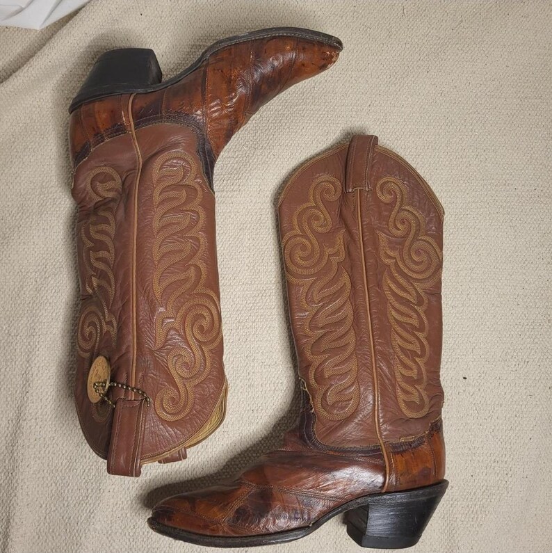 May include: A pair of brown leather cowboy boots with intricate stitched designs. The boots have pointed toes, stacked heels, and decorative stitching. One boot has a tag attached. The boots are a medium brown color.