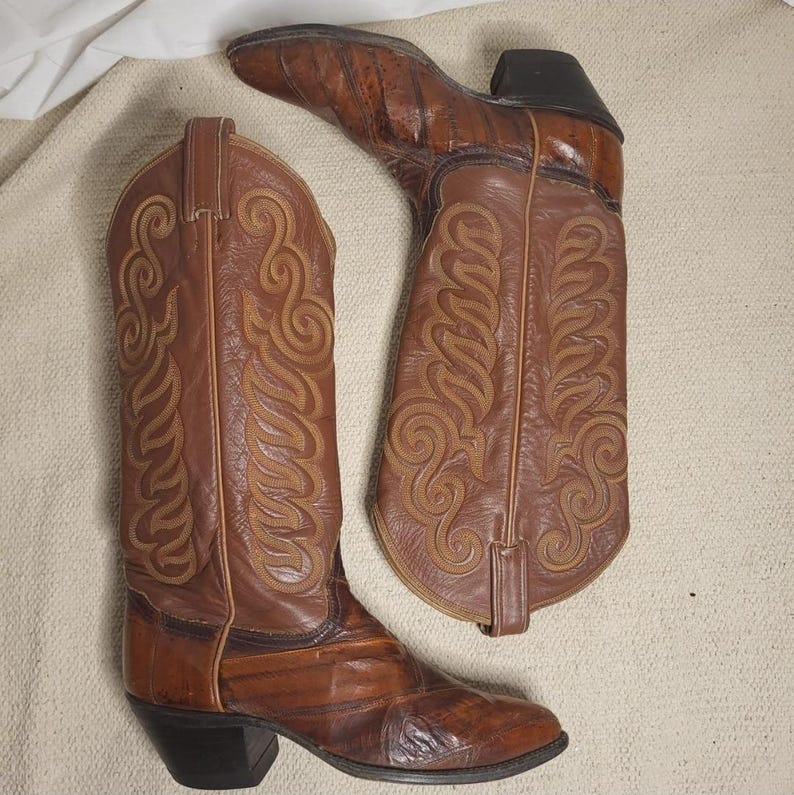 May include: A pair of brown leather cowboy boots with intricate tan stitching. The boots have pointed toes, stacked heels, and decorative patterns on the shafts. The boots are a classic Western style.