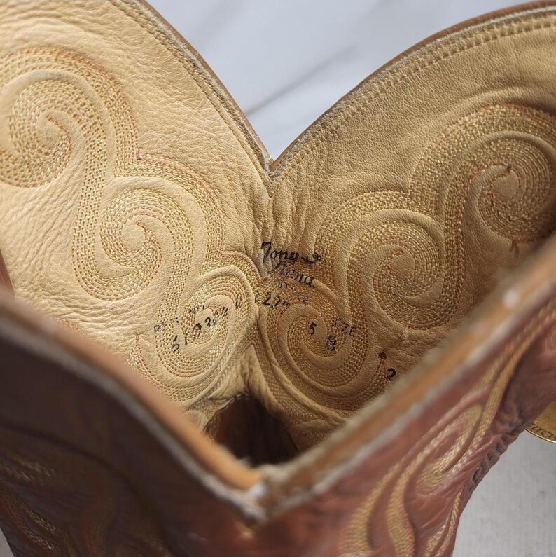 May include: Close-up of the interior of a brown leather cowboy boot. The boot's interior features a tan leather lining with an embossed swirl design. The brand name "Tony Lama" is visible, along with size information.