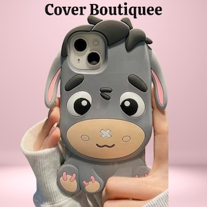 May include: A grey phone case with a cartoon donkey design. The case has large eyes, pink-lined ears, and a stitched-mouth detail. The text "Cover Boutiquee" is at the top. The case is held against a pink background.