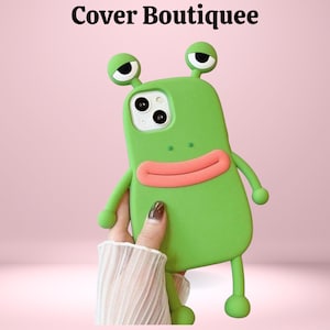 Puede incluir: Funda de teléfono verde con un diseño de rana de dibujos animados. La funda tiene un cuerpo verde claro, una boca rosa y dos ojos grandes y redondos. La funda tiene brazos y piernas, y el texto "Cover Boutiquee" está en la parte superior.