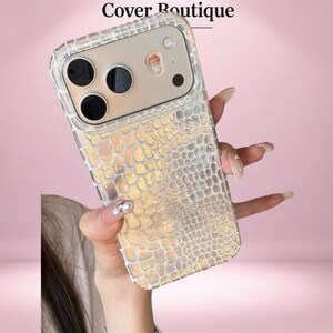 Puede incluir: Una funda transparente para smartphone con un patrón de piel de cocodrilo texturizado en tono dorado. La funda incluye un recorte para las lentes de la cámara y un pequeño dije decorativo circular. El texto Cover Boutique aparece en la parte superior.