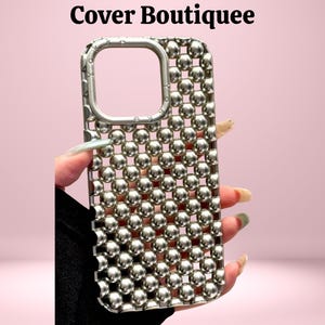Puede incluir: Una funda plateada para teléfono con una rejilla de pequeñas esferas brillantes. La funda tiene un recorte cuadrado para la cámara y el texto "Cover Boutiquee" en la parte superior. La funda se sostiene sobre un fondo rosa.