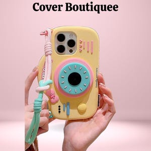 Op de afbeelding: Gele telefoonhoes met een camera-ontwerp, met een roze en blauwe cirkelvormige lens en een bijpassende polsband. De hoes heeft een speelse uitstraling, met de tekst "Lovely Camera" aan de zijkant.