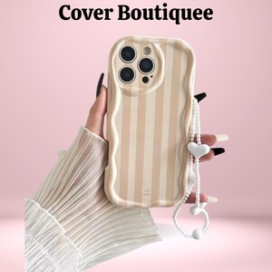Könnte beinhalten: Beige und weiß gestreifte Handyhülle mit gewelltem Rand und weißem herzförmigem Charmanhänger. Die Handyhülle hat drei Kameraausschnitte und den Text "Cover Boutiquee" oben.