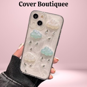 Puede incluir: Funda transparente para teléfono con fondo nacarado blanco, adornada con adornos en forma de nube azul claro y blanco y brillantes pedrería en forma de lágrima. El texto "Cover Boutiquee" está en la parte superior.