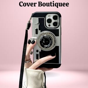 May include: Black and silver camera-style phone case with a black strap. The case has a camera lens design and the text "Cover Boutiquee" at the top. The phone case is held against a pink background.