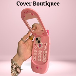 Può includere: Custodia rosa per telefono a conchiglia con specchio, dettagli a cuore e a stella. La custodia ha una tastiera con numeri e una piccola antenna. Il testo "Cover Boutiquee" è in alto.