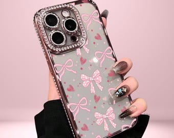 Pode incluir: Capa de telefone transparente com um padrão de laço e coração rosa. A capa tem uma moldura prateada com uma borda de strass ao redor da câmera. O texto "Cover Boutiquee" está no topo.