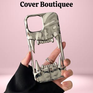May include: A silver-colored phone case with a fang design. The case has a glossy finish and features sharp, pointed fangs and teeth. The text "Cover Boutiquee" is at the top of the image. The case is held against a pink background.