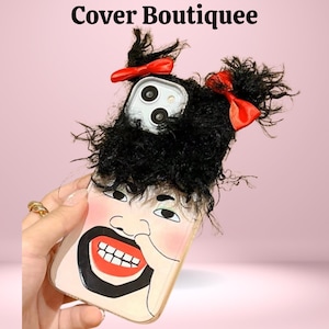 May include: A novelty phone case featuring a cartoon face design with black hair, red bows, and a smiling mouth. The case has a unique, textured appearance and the text "Cover Boutiquee" is visible at the top.