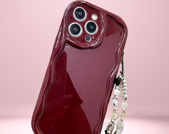May include: A burgundy phone case with a wavy edge design and a decorative beaded wrist strap. The case has a glossy finish and features the text "Cover Boutiquee" at the top. The strap includes clear, white, and black beads.