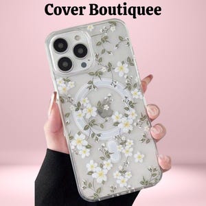 May include: Clear phone case with a floral design featuring white flowers and green leaves. The case has a magnetic ring and the text "Cover Boutiquee" at the top. The case is held against a pink background.