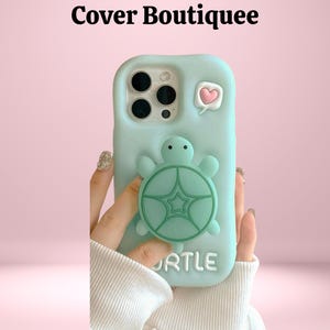 Puede incluir: Funda de teléfono azul claro con un diseño de tortuga en 3D. La funda tiene un bocadillo en forma de corazón y la palabra "URTLE" en la parte inferior. La funda se sostiene sobre un fondo rosa.