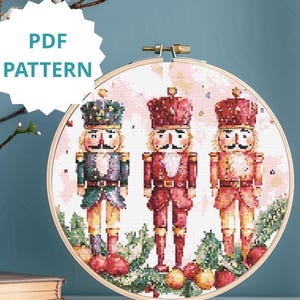 May include: A cross-stitch pattern featuring three toy soldier nutcrackers in a wooden embroidery hoop. The nutcrackers are dressed in red, green, and gold uniforms, with crowns and mustaches. The text "PDF PATTERN" is in a white starburst.
