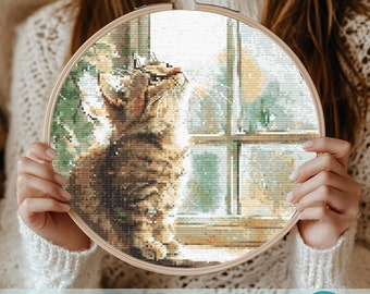 May include: A cross stitch pattern featuring a brown tabby cat looking out a window. The cat is sitting on a windowsill with a view of a snowy landscape. The pattern is in a wooden embroidery hoop.