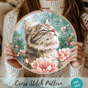 May include: Cross stitch pattern featuring a cat with closed eyes and pink flowers. The cat is surrounded by green leaves and pink flowers. The pattern is in a wooden embroidery hoop.
