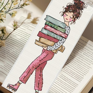 May include: A rectangular cross-stitch bookmark featuring a person with a high bun hairstyle carrying a stack of five books. The person wears a gray patterned long-sleeve shirt and pink trousers with matching pink shoes, rendered in a grid-like stitch pattern.