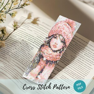 Pode incluir: Padrão de ponto cruz para um marcador de página com uma mulher usando um chapéu e um cachecol rosa. O padrão está em formato PDF.