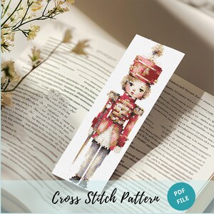 Puede incluir: Un patrón de punto de cruz con una ilustración colorida de un soldado de juguete. El soldado lleva un uniforme rojo y dorado con un sombrero blanco y pantalones blancos. El patrón está impreso en papel blanco e incluye el texto "Cross Stitch Pattern" y "PDF FILE".