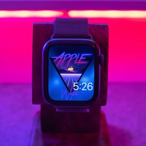 Könnte beinhalten: Eine schwarze Apple Watch mit einem dunkelgrauen Metallband befindet sich auf einem hölzernen Ladeständer. Das Zifferblatt der Uhr ist ein blaues und pinkfarbenes Retro-Design mit dem Text "Apple Watch" und der Zeit 5:26.