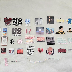 May include: A collection of various stickers with diverse designs and text. The stickers feature band logos, phrases like "No Me," and images of musicians. Some stickers have text like "The Clancy World Tour" and "Peace, Love & Jim Dun."