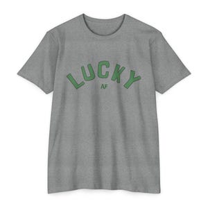 Könnte beinhalten: Ein hellgraues T-Shirt mit einer grünen Grafik, auf der in einer Retro-Schrift "LUCKY AF" steht.