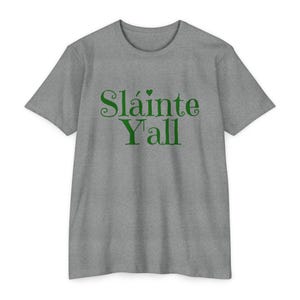 May include: A heather gray t-shirt with the green text "Sláinte Y'all". The text is in a decorative font with a small green heart above the "i". The shirt has short sleeves and a crew neck.