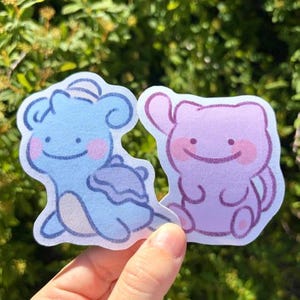 May include: Two cartoon stickers featuring a blue and a pink character with smiling faces and pink cheeks. The blue character has a shell-like design, while the pink character has a simple, rounded shape. The stickers are held against a green, leafy background.