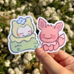 May include: Two colourful stickers featuring cartoon characters. One sticker depicts a yellow and green character with a blue flower, and the other is a pink fox-like character. The stickers are held by a hand against a background of white flowers.