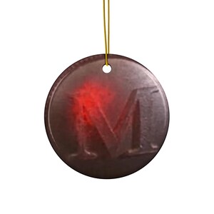 Op de afbeelding: Een cirkelvormig, donkerkleurig ornament met een opvallende, in reliëf aangebrachte letter M in het midden. Het oppervlak heeft een metalen, getextureerde uitstraling met een heldere rode gloed die uit de linkerkant van de letter komt.