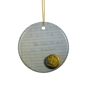 Può includere: Un ornamento rotondo in ceramica bianca con un cordino dorato per appenderlo. L'ornamento presenta una nota scritta a mano con il testo "Be Sure To Drink Your. OVALTINE" e un bottone dorato.