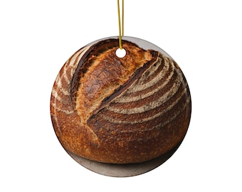May include: Round ceramic ornament featuring a detailed image of a loaf of artisan bread. The bread has a dark brown crust with lighter brown scoring and a golden hanging cord. The ornament is round and has a hole at the top for hanging.