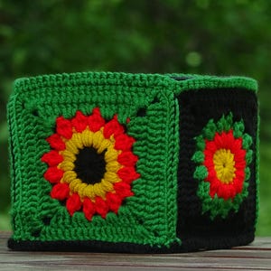 May include: A handmade, square crochet box featuring a vibrant floral design. The box is primarily green with black accents and colorful flowers in red, yellow, and black. The crochet stitches are clearly visible, showcasing the handcrafted nature of the item.