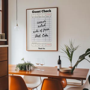 May include: A framed print of a vintage guest check with the handwritten order "Caesar salad, side of fries, Diet Coke" and the number 178562. The print is hanging on a white wall above a wooden table with two wine glasses, a bottle of wine, and a plant.