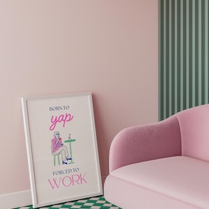 以下が含まれることがあります： ピンクとブルーで「BORN TO yap FORCED TO WORK」の文字が入った額入りのアートプリント。テーブルに座っている人の漫画イラストが特徴です。部屋にはピンクの壁、緑と白の市松模様の床、ピンクのアームチェアがあります。
