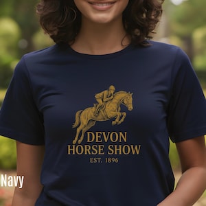 May include: Navy blue t-shirt with a gold illustration of a horse and rider jumping. The text "DEVON HORSE SHOW EST. 1896" is printed below the image. The word "Navy" is printed in white at the bottom left of the t-shirt.