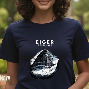 Puede incluir: Camiseta azul marino con un gráfico de la cara norte del Eiger. El texto "EIGER NORTH FACE" está impreso encima de la ilustración de la montaña. El gráfico de la montaña es en blanco y negro.
