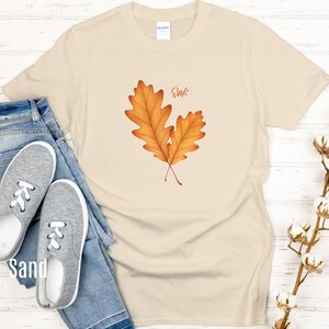May include: Sand-colored t-shirt featuring a watercolor illustration of two oak leaves in shades of orange and brown. The word "Oak" is written above the leaves in a handwritten font. The shirt is paired with blue jeans and gray sneakers.
