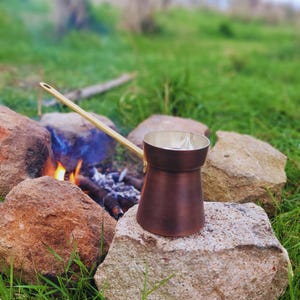 Op de afbeelding: Een koperkleurige koffiepot met een lange, goudkleurige handgreep staat op een grote rots. Er is een klein kampvuur met vlammen en sintels in de buurt. De achtergrond toont groen gras en rotsen, wat wijst op een buitenomgeving.