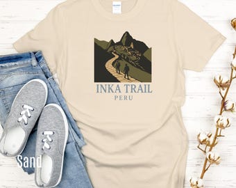 Puede incluir: Camiseta color arena con un gráfico del Camino Inca en Perú. El diseño incluye un camino que conduce a una montaña con ruinas antiguas y el texto "INKA TRAIL PERU". La camiseta se combina con vaqueros azules y zapatillas grises.
