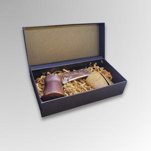 Puede incluir: Una caja de regalo azul oscuro con interior marrón, llena de papel triturado y varios artículos. Se ve un objeto cilíndrico marrón, un tubo plateado y un artículo de color beige. La caja está abierta, revelando el contenido.