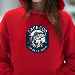 Può includere: Una felpa con cappuccio rossa con una grafica circolare raffigurante un faro, un pesce che salta e le parole "CAPE COD STRIPER FISHING" in bianco e blu navy. La parola "Red" è scritta in corsivo in basso a destra.